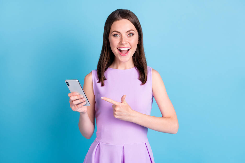 Fotografie užaslý šťastný veselá mladá žena ukazováček držet chytrý telefon izolovat na lesk modré barvy pozadí - Fotografie, Obrázek