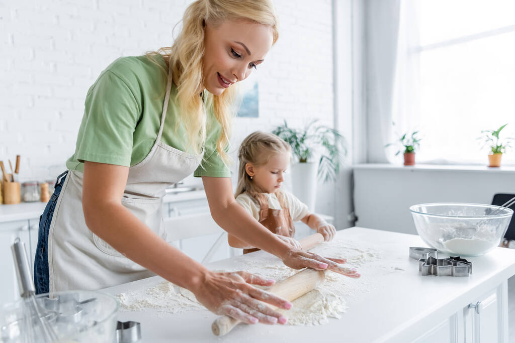 Frau mit kleiner Tochter rollt rohen Teig auf Küchentisch - Foto, Bild