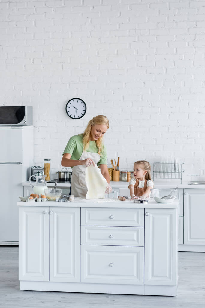 sonriente mujer sosteniendo la masa cerca de su hijita en la cocina - Foto, imagen