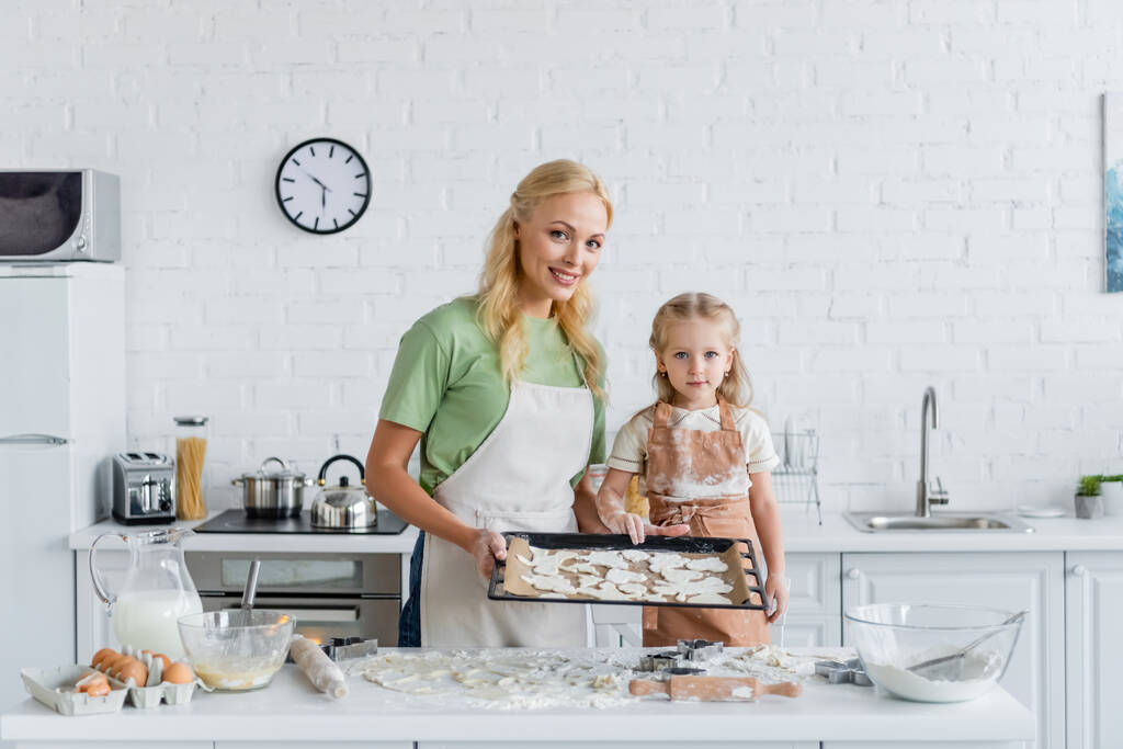 счастливые мама и ребенок с сырым печеньем на хлебопекарном листе, глядя на камеру на кухне - Фото, изображение