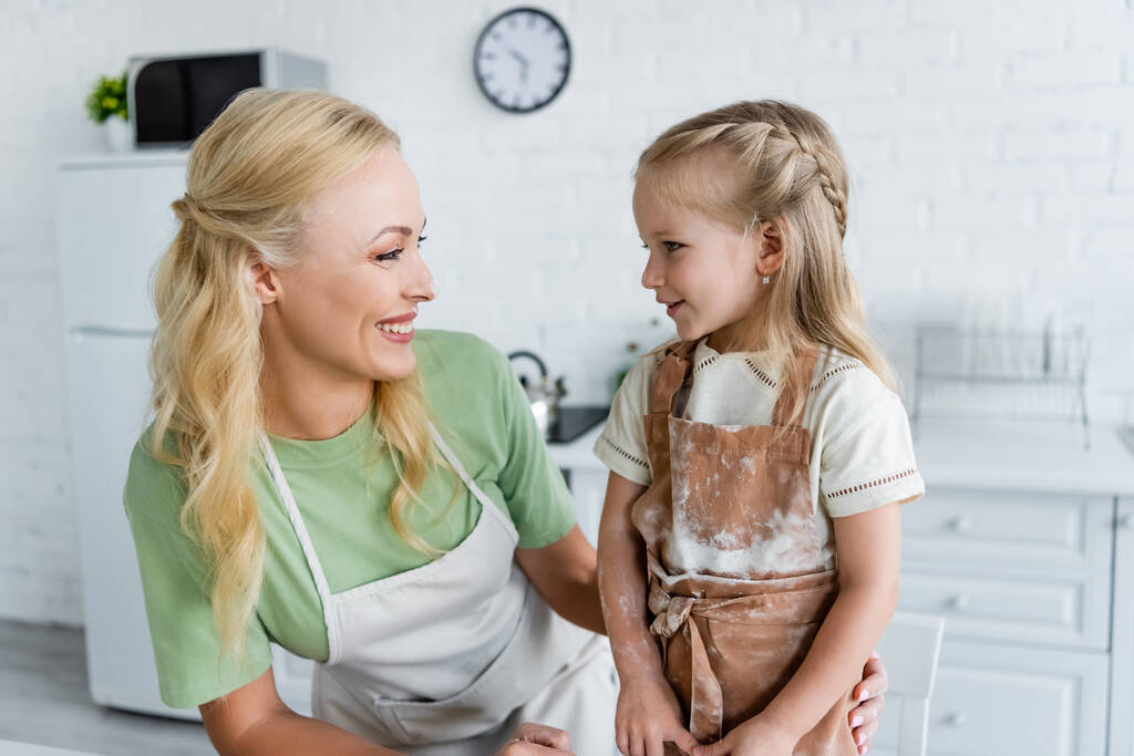 mujer feliz mirando a la hija en delantal con manchas de harina en la cocina - Foto, Imagen