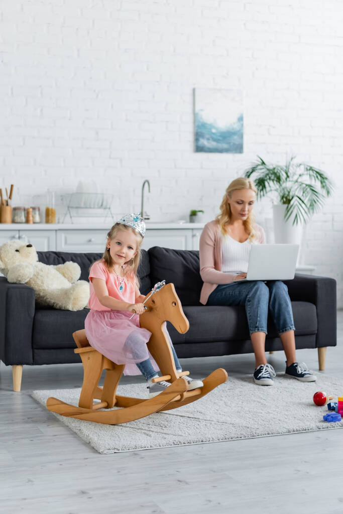 ребенок в игрушечной короне верхом на качающейся лошади рядом с размытой мамой работает на ноутбуке - Фото, изображение