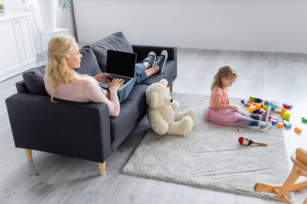 γυναίκα δακτυλογράφηση σε φορητό υπολογιστή στον καναπέ κοντά κόρη παίζει με δομικά στοιχεία στο πάτωμα - Φωτογραφία, εικόνα