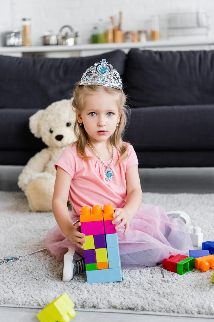 Девушка в игрушечной короне смотрит в камеру рядом с башней красочных строительных блоков - Фото, изображение