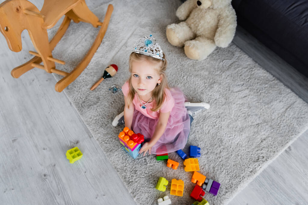Blick von oben auf Kind in Spielzeugkrone, das mit bunten Bausteinen in der Nähe von Spielzeug auf dem Boden spielt - Foto, Bild