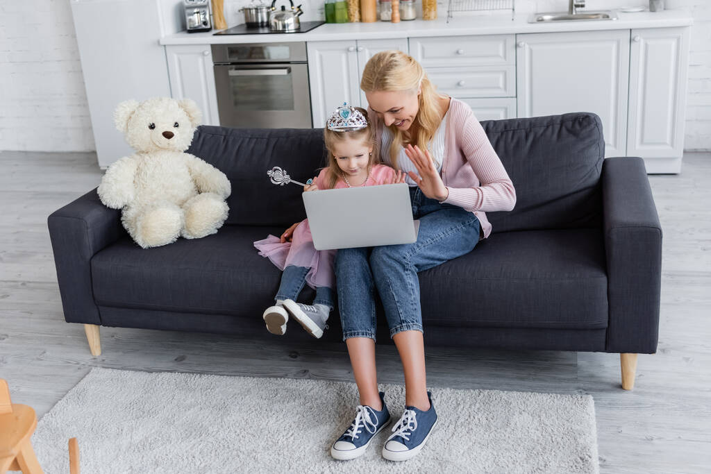 onnellinen nainen tyttärensä kanssa lelu kruunu heiluttaa käsiä videopuhelun aikana kotona - Valokuva, kuva