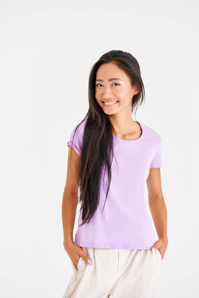 Довольно улыбающаяся азиатка в повседневной одежде, позирующая перед камерой на белом фоне - Фото, изображение