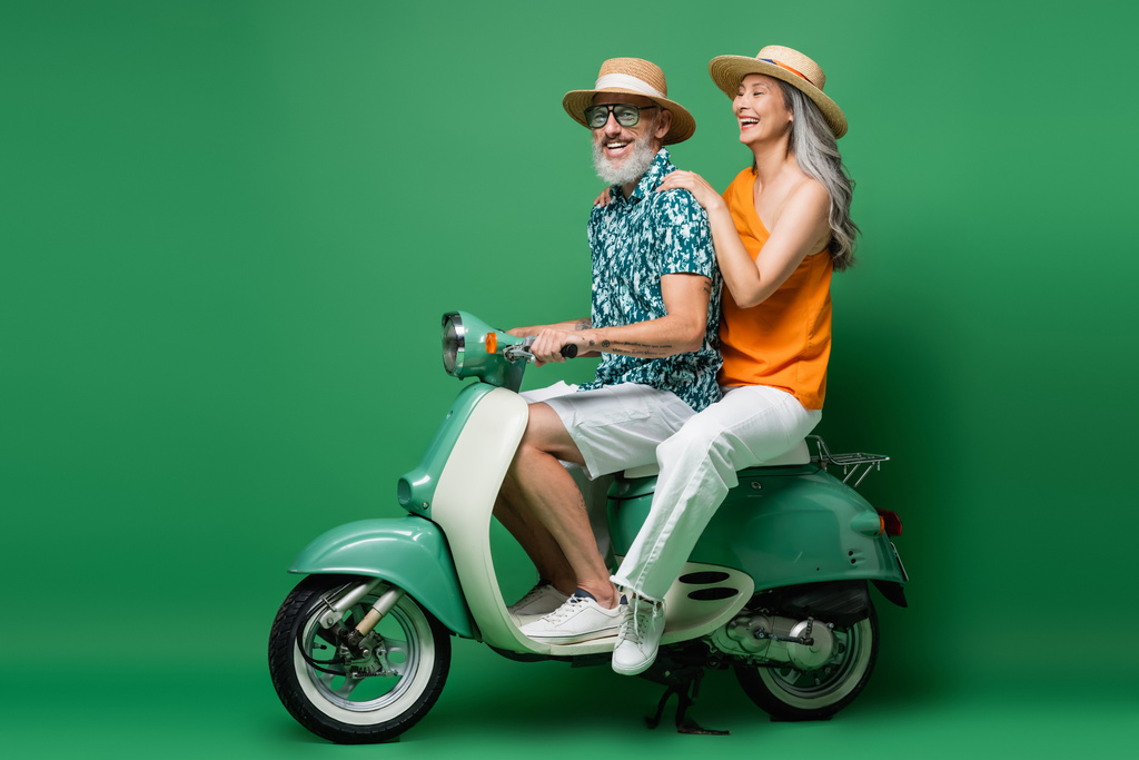 веселая межрасовая пара средних лет в солнечных шляпах катается на мопеде на зеленом  - Фото, изображение