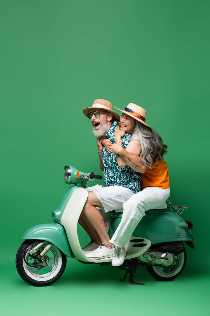 возбужденная и межрасовая пара средних лет в солнечных шляпах обнимается во время езды на мотороллере на зеленом - Фото, изображение