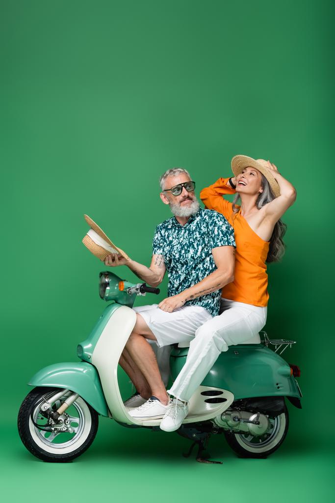 ευτυχισμένη και πολυεθνική μεσήλικη γυναίκα κρατώντας καπέλο ήλιο, ενώ ιππασία μοτοποδήλατο με το σύζυγο στο πράσινο  - Φωτογραφία, εικόνα