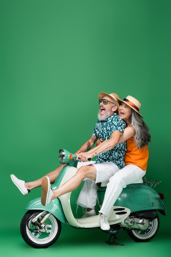 испуганная и многонациональная пара средних лет в солнечных шляпах, катающаяся на мопеде на зеленом - Фото, изображение