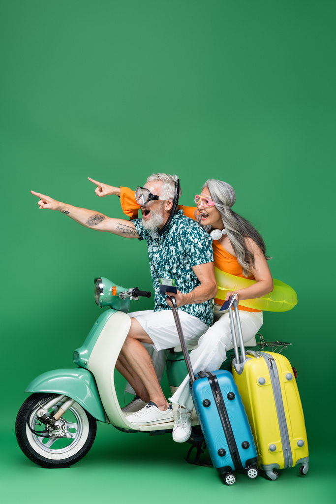 изумленная и многонациональная пара средних лет в очках, указывающих пальцами и держащих паспорта во время езды на мотороллере на зеленом - Фото, изображение