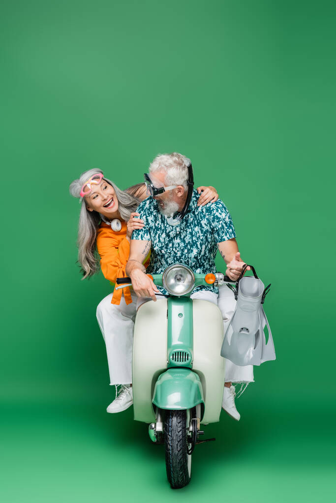 fröhliche asiatische Frau mittleren Alters, die ihren Mann in Brille mit Schwimmflossen ansieht, während sie Motorroller auf Grün fährt  - Foto, Bild
