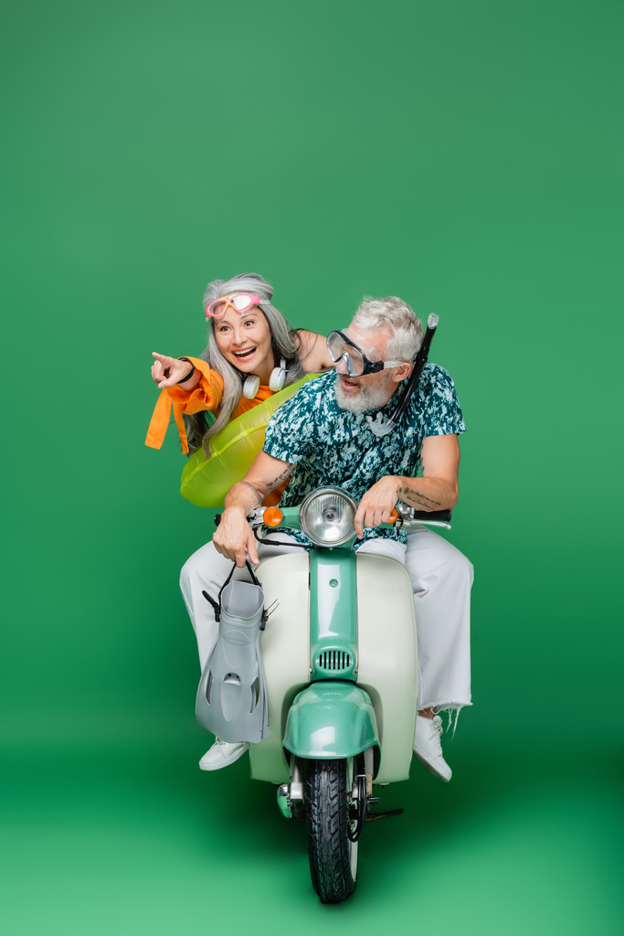 весела азіатська жінка середнього віку вказує пальцем біля чоловіка тримає лапки під час їзди на моторолері на зеленому
  - Фото, зображення