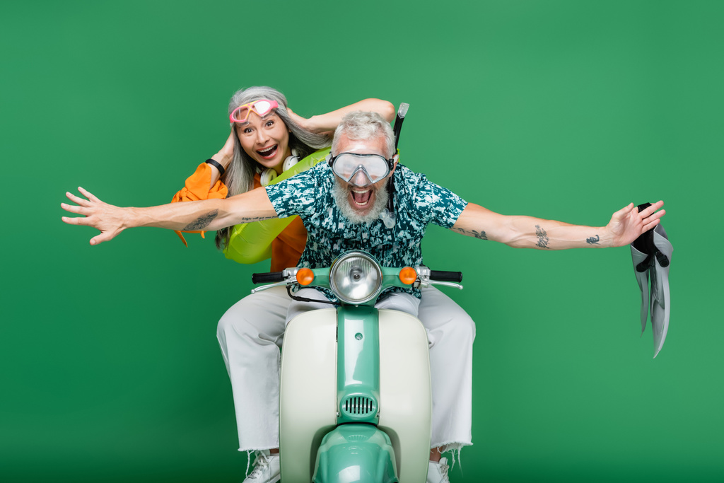 cheerful middle aged man with outstretched hands holding flippers near asian wife in goggles riding moped on green  - Photo, Image