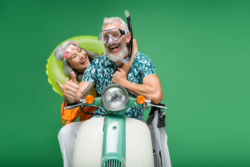 cheerful middle aged asian woman hugging husband in goggles showing thumb up while riding motor scooter isolated on green  - Photo, Image