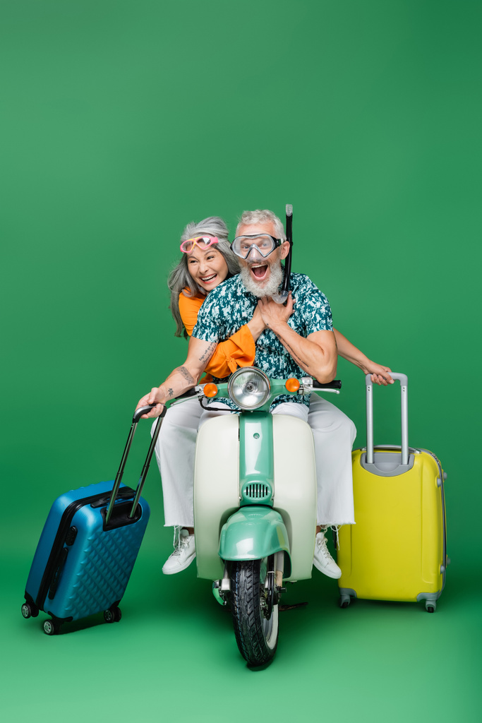amazed and mature multiethnic couple in goggles holding luggage while riding moped on green - Photo, Image