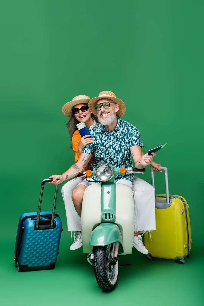 alegre casal de meia-idade e multiétnico em chapéus de sol segurando passaportes e bagagem enquanto montava ciclomotor em verde - Foto, Imagem