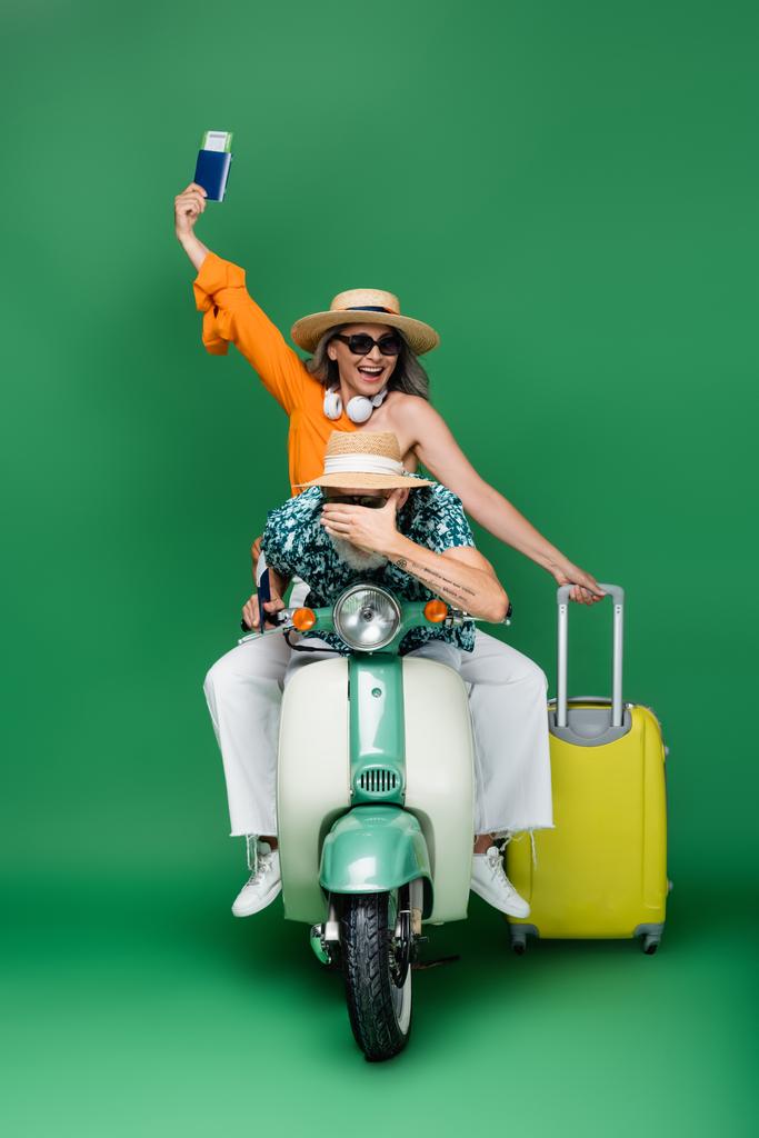 wesoły średnim wieku Azji kobieta w sunhat gospodarstwa paszport w pobliżu męża obejmujące oczy podczas jazdy skuter motorowy na zielony - Zdjęcie, obraz