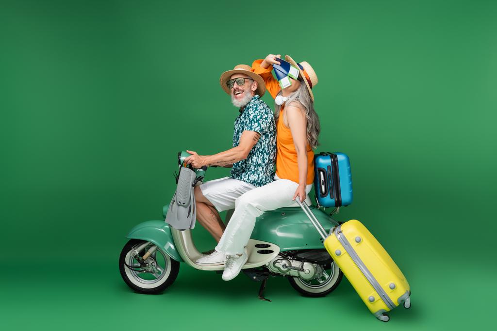 maduro asiático mujer en sol sombrero cubriendo cara con pasaportes cerca marido celebración aletas mientras cabalgando ciclomotor en verde - Foto, imagen