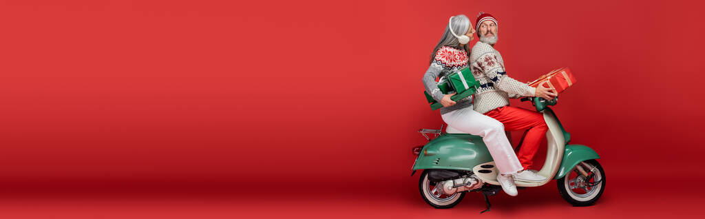 Χαρούμενη μεσήλικη και Ασιάτισσα με ωτοασπίδες που κρατάνε χριστουγεννιάτικα δώρα ενώ καβαλάνε μοτοποδήλατο με τον σύζυγο πάνω σε κόκκινο πανό - Φωτογραφία, εικόνα