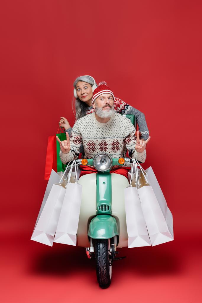 hombre de mediana edad mostrando señal de roca cerca sorprendido esposa asiática con bolsas de compras mientras monta moto en rojo - Foto, Imagen