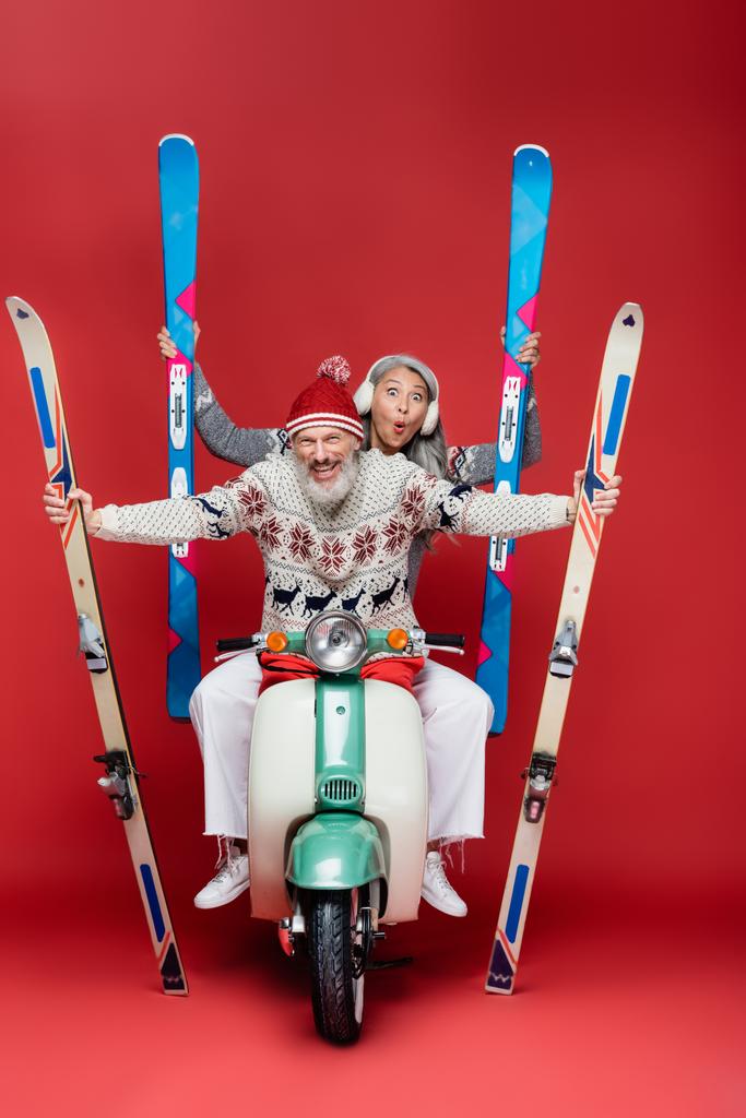 cheerful interracial and middle aged couple in sweaters holding skis while riding motor scooter on red - Photo, Image