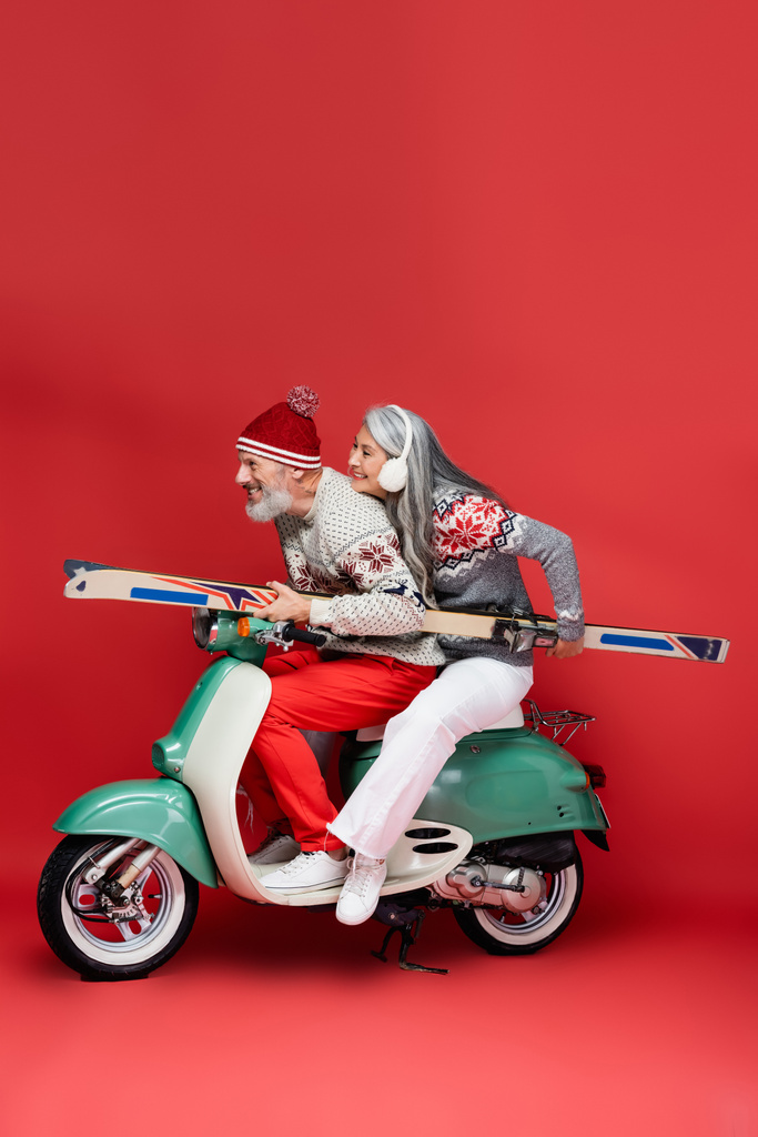 widok z boku szczęśliwy międzyrasowy i w średnim wieku para w swetrach gospodarstwa narty podczas jazdy motorower na czerwonym - Zdjęcie, obraz