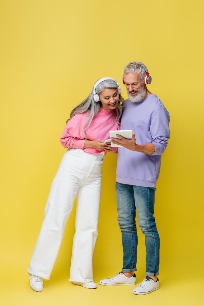 黄色のガジェットを使用してワイヤレスヘッドフォンで幸せな中年と異人種間のカップルの完全な長さ - 写真・画像