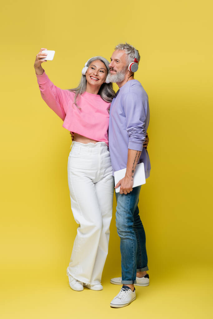 full length of happy interracial and middle aged couple in wireless headphones taking selfie on yellow - Photo, Image