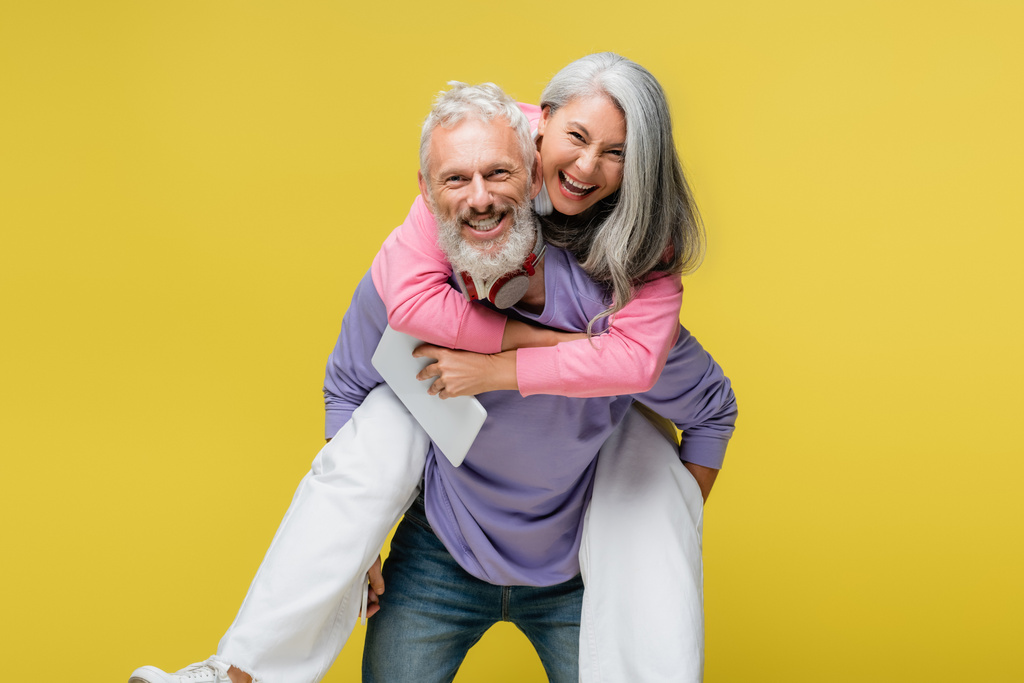 cheerful middle aged man piggybacking happy asian wife with digital tablet isolated on yellow  - Photo, Image