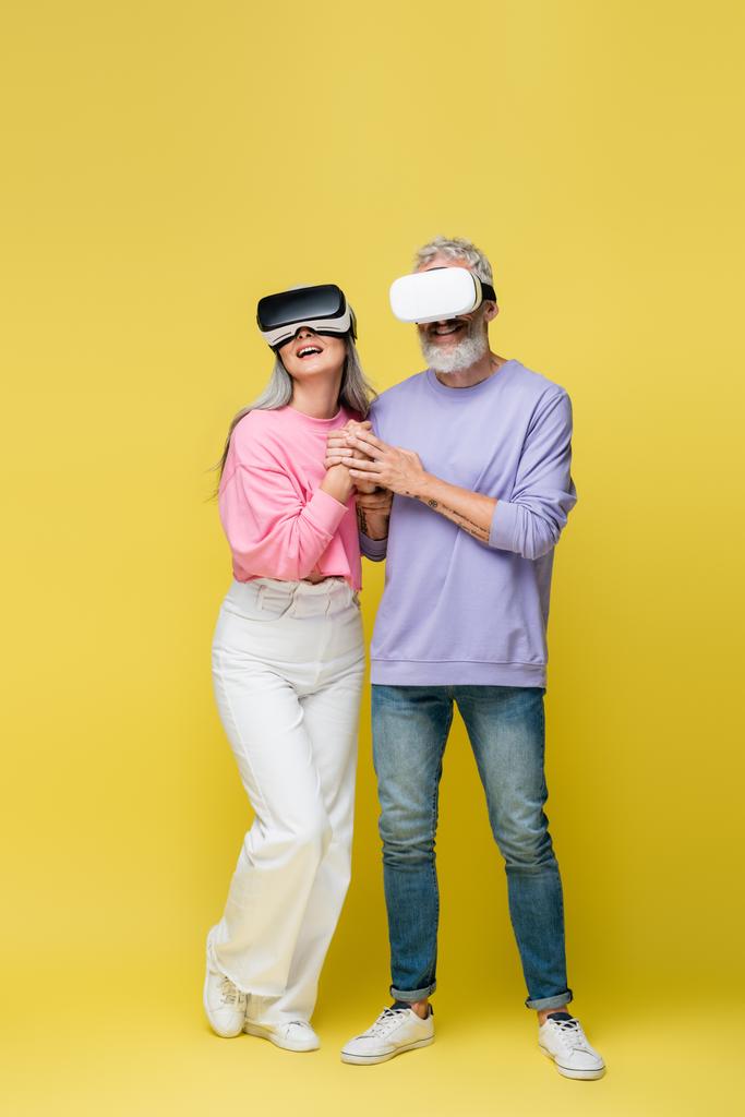 full length of multiethnic and middle aged couple in vr headsets holding hands while gaming on yellow - Photo, Image