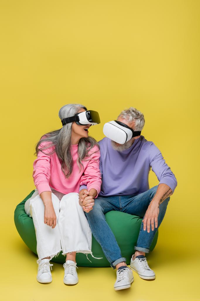 interracial and cheerful middle aged couple in vr headsets sitting in bean bag chair on yellow  - Photo, Image