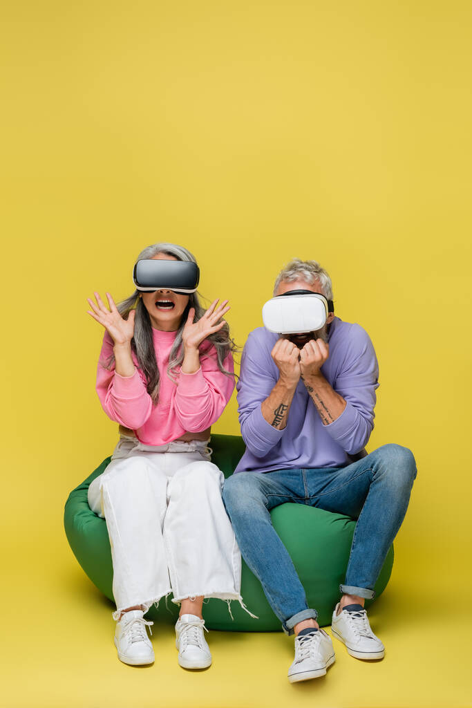 interrassische und ängstliche Paar mittleren Alters in vr Headsets sitzt in Bean Bag Stuhl, während Gaming auf gelb  - Foto, Bild