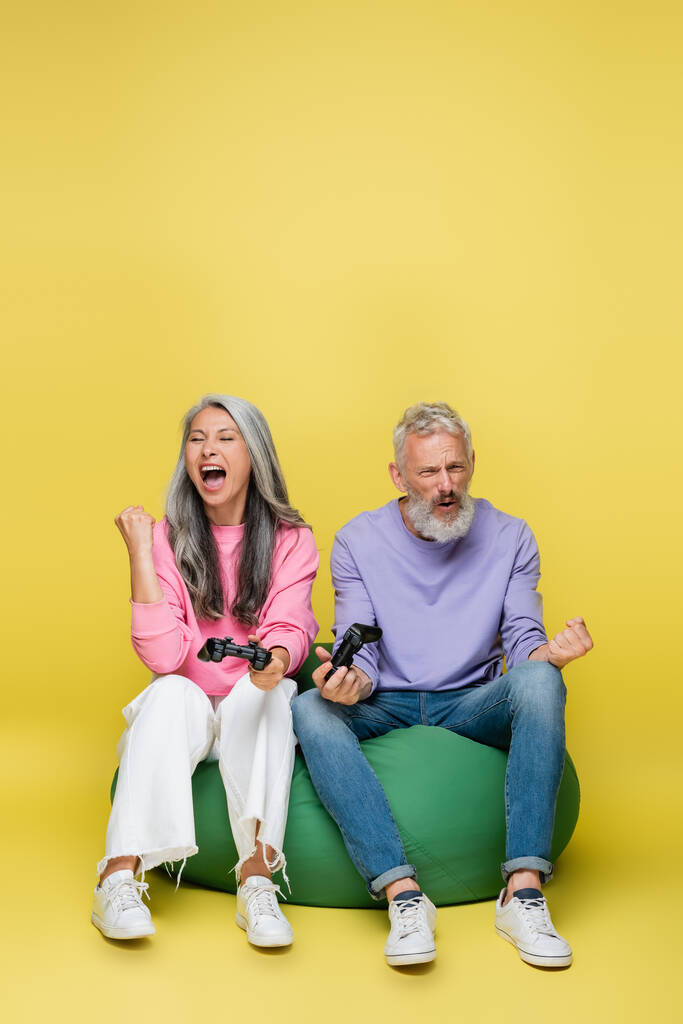 KYIV, UKRAINE - 10 AOÛT 2021 : couple d'âge moyen interracial et excité tenant des joysticks après avoir gagné un jeu vidéo sur jaune  - Photo, image