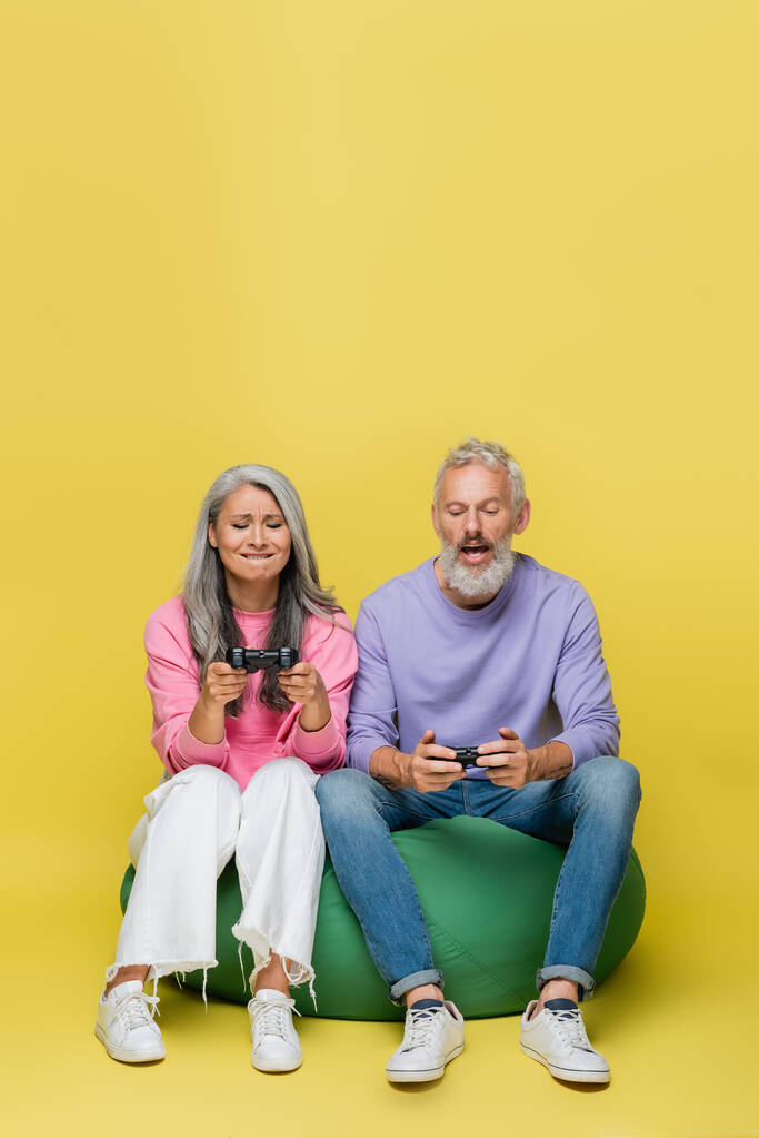 KYIV, UCRANIA - 10 de agosto de 2021: pareja interracial de mediana edad tensa sosteniendo joysticks y jugando videojuegos en amarillo  - Foto, Imagen