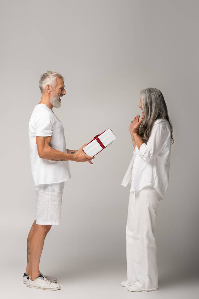 volledige lengte van gelukkig middelbare leeftijd man geven gewikkeld gift box naar verbaasd aziatische vrouw op grijs - Foto, afbeelding