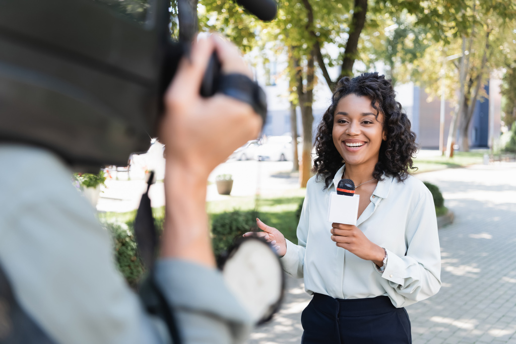 cámara borrosa con cámara de video cerca sonriente reportero afroamericano con micrófono haciendo reportaje - Foto, Imagen