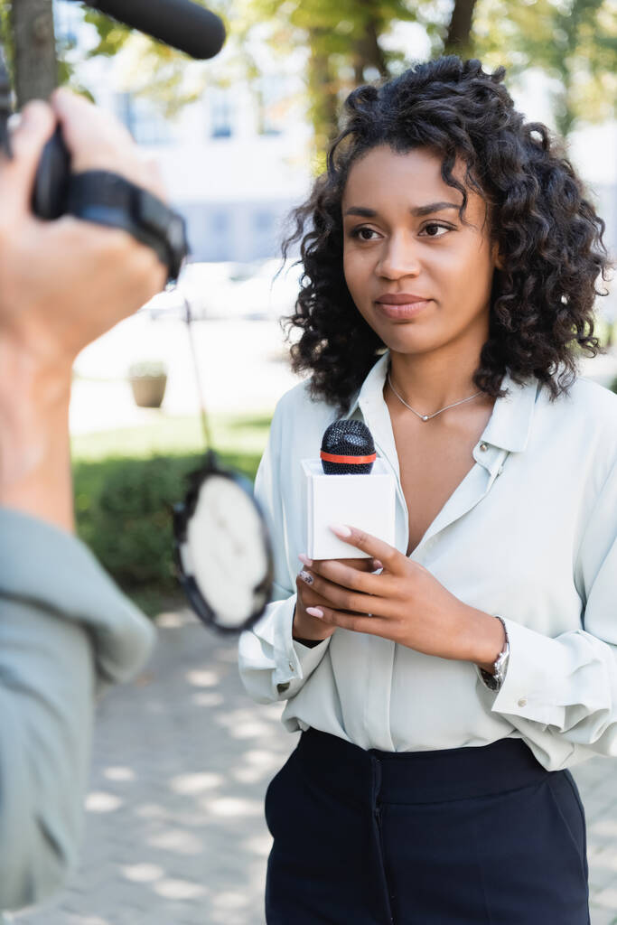 όμορφη αφροαμερικανή δημοσιογράφος με μικρόφωνο κάνει ρεπορτάζ κοντά θολή κάμεραμαν με βιντεοκάμερα  - Φωτογραφία, εικόνα