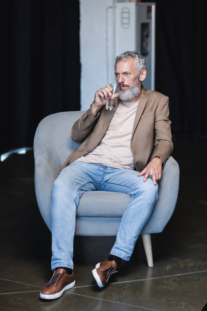 szakállas üzletember kezében pohár víz és ül szürke fotelben interjú közben - Fotó, kép
