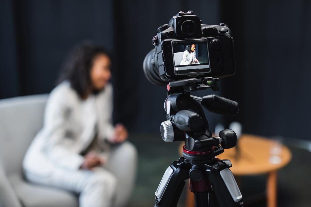 современная цифровая камера на штативе с африканским американским журналистом, сидящим в кресле на экране  - Фото, изображение