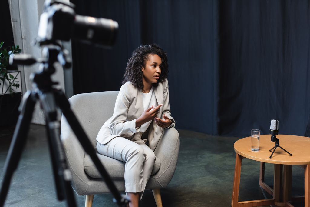 afro-amerikai újságíró ül karosszékben, és beszél közelében elmosódott digitális fényképezőgép állvány - Fotó, kép