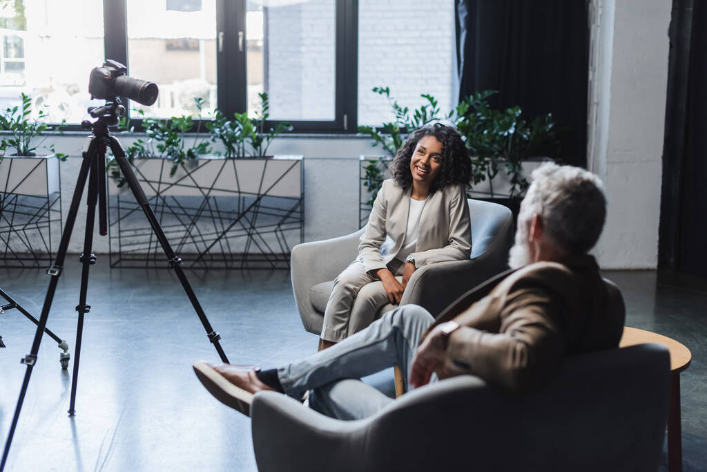 щасливий афроамериканський журналіст і розмитий бізнесмен розмовляє під час інтерв'ю біля цифрової камери на тринозі
 - Фото, зображення