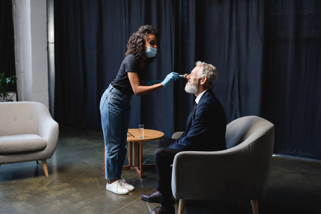 curly african american makeup artist in medical mask and latex gloves applying face powder on face of businessman  - Photo, Image
