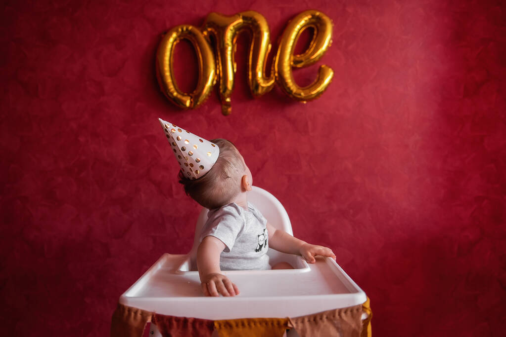 Kleine jongen van een jaar in feestelijke hoed zit in kinderstoel met stof vlaggen, kijkt naar de folie gouden ballonnen Een. Kind op rode geïsoleerde achtergrond, op getextureerde muur vieren vakantie thuis - Foto, afbeelding