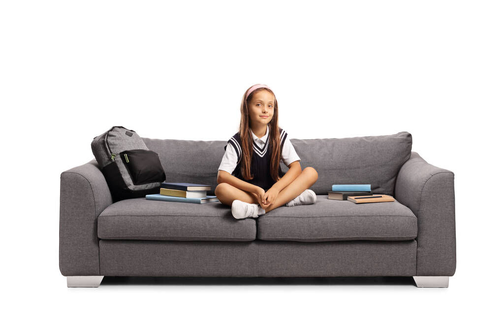 Schulmädchen in Uniform sitzt auf einem Sofa mit Büchern und einem Rucksack auf weißem Hintergrund - Foto, Bild