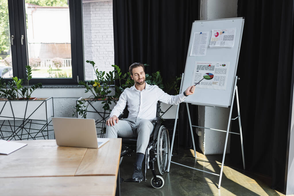 disabled businessman in wheelchair pointing at flip chart during video call on laptop in office - Photo, Image