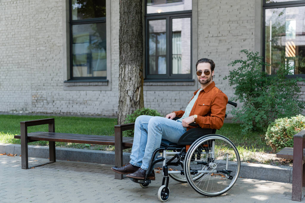 jeune homme handicapé en fauteuil roulant souriant à la caméra dans la rue urbaine - Photo, image