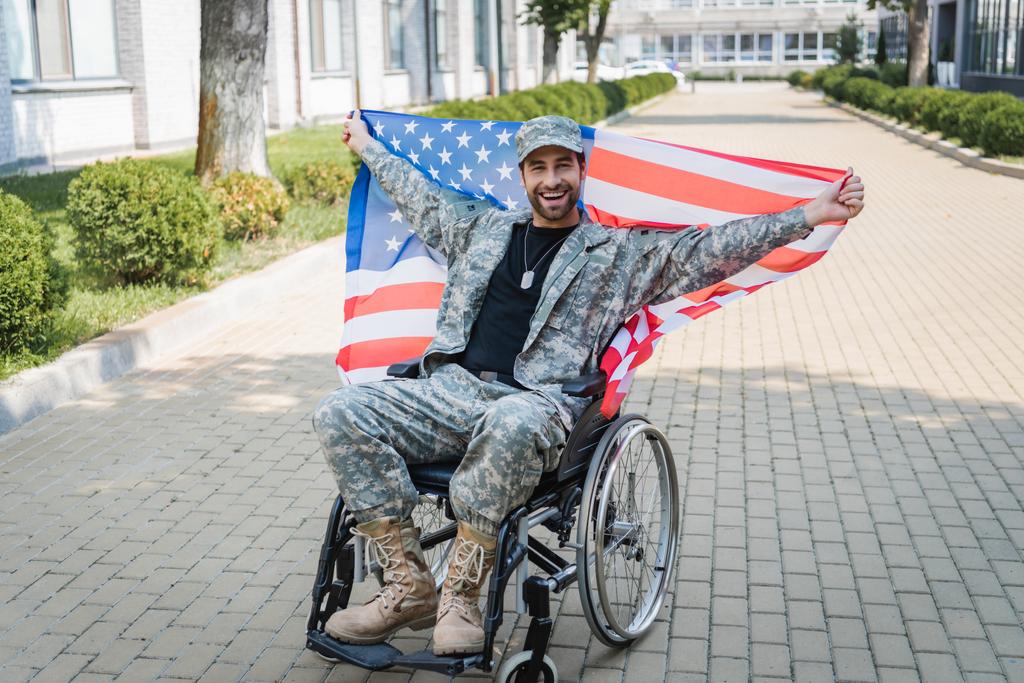 ευτυχής βετεράνος με αναπηρία κοιτάζοντας κάμερα, ενώ κάθεται σε αναπηρική καρέκλα με σημαία ΗΠΑ - Φωτογραφία, εικόνα