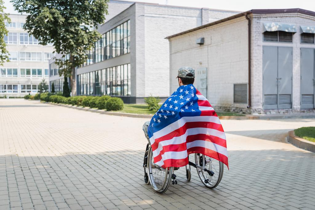 back view of young veteran, covered with usa flag, sitting in wheelchair in city - Photo, Image